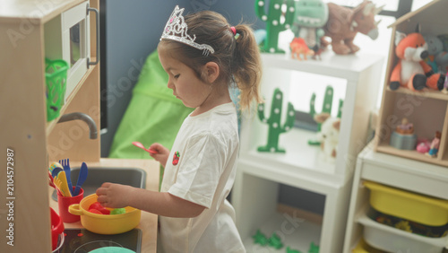Fotografía Little girl wearing tiara stirs plastic bowl with red spoon at toy sink in a playroom building; playful imagination