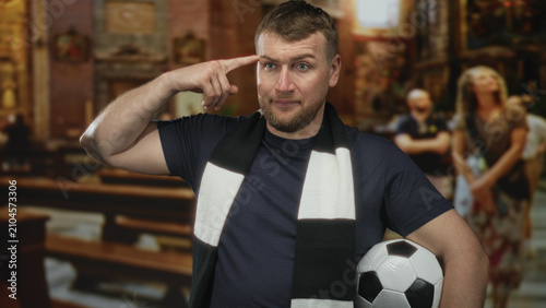 Cuadro en lienzo Man with soccer ball and black and white fan scarf points finger to temple inside a church building while leaning on a pew and glancing sideways; mischievous irreverence conflict