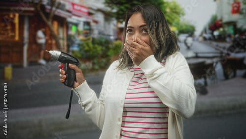 Cuadro en lienzo Hispanic middle aged woman holding a cordless screwdriver, hand covering mouth gesture, looking startled on a busy street with storefronts; surprise concern