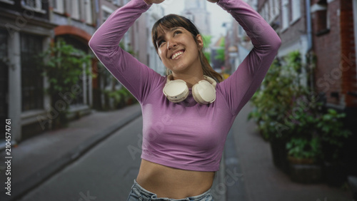 Foto Woman wearing headphones raises arms overhead, midriff visible on a street with purple top and jeans; joyful spontaneity