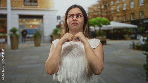 Canvas Print Young caucasian woman with hands clasped to chest on city street wearing red glasses and white top; frustration