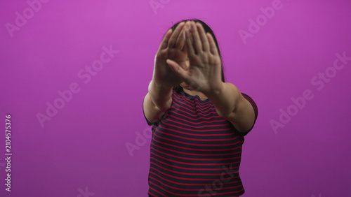 Photography Woman with palms forward covering face in a purple studio, arms extended toward camera wearing red and navy striped shirt; privacy defensive