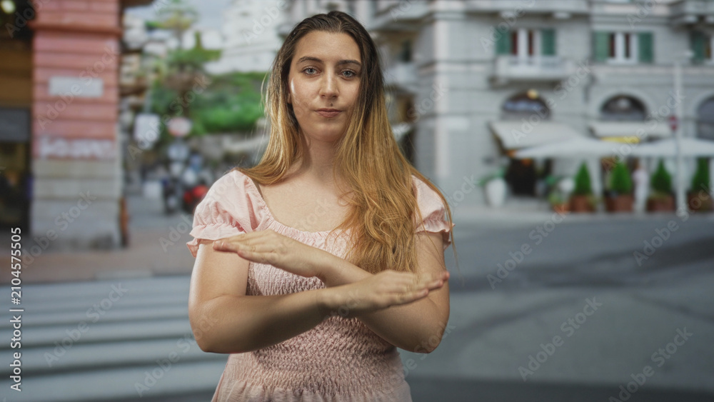 Fototapeta premium Woman crosses arms in an x over her chest with hands visible on a street crosswalk by sidewalk cafe umbrellas, wearing pink dress; defiance refusal.