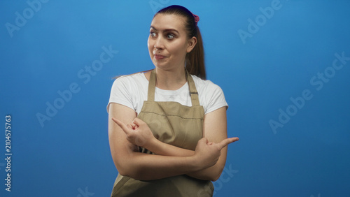 фотографія Woman in apron and white tshirt points both index fingers crosswise while smiling toward side in studio; playfulness humor