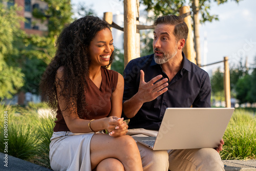 Two diverse business people gathered for a briefing outside the office working with a laptop, young woman and middle age man collaborating together