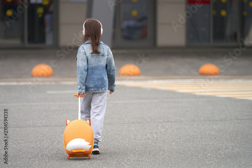 A young school-age girl wearing headphones approaches the airport terminal, pulling a small suitcase on wheels.