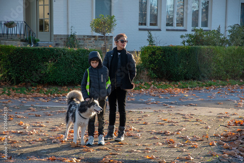 Woman and boy enjoying an outdoor walk with their pet husky dog on a street covered with fallen autumn leaves, showing family bond and leisure activity