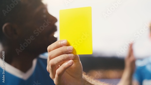 Tableau sur toile A close-up shows a referee holding a yellow card after a rules violation during a soccer match, emphasizing the official warning, disciplinary decision and sanction for the foul on the pitch