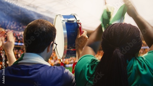 Obraz na plátně Man beats a drum inside a football stadium during match day while a woman chants beside him, creating crowd rhythm and motivating players as supporters cheer from the stands