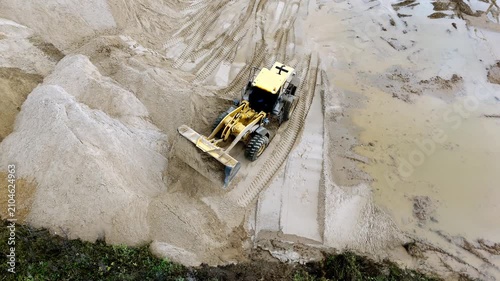 Aerial view shows a bulldozer moving sand in a quarry. Yellow equipment pushes material through muddy tracks. Large sand piles form dry terrain