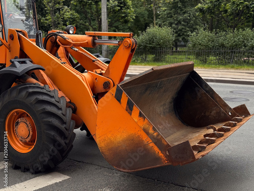 Close up view of an orange front wheel loader bucket and tire on a city asphalt road. Heavy industrial machinery equipment. Large metal bucket of an orange excavator vehicle on the street. 