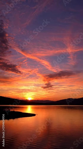 Magnificent sunset paints the sky in vibrant hues of orange, purple, and gold over a tranquil lake, reflecting the dramatic clouds