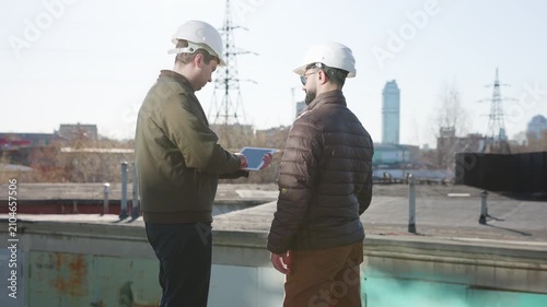 Two construction workers in hard hats shaking hands on rooftop during urban site review with power lines, buildings, and industrial skyline in background, with construction, worker, hardhat, rooftop