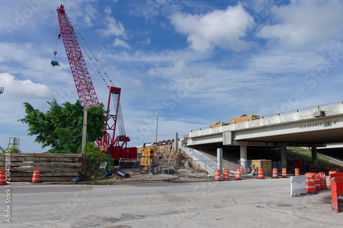 Red crane lifts materials beside a concrete highway overpass surrounded by orange traffic barrels, construction equipment and excavated earth in St. Petersburg,FL illustrating active infrastructure re