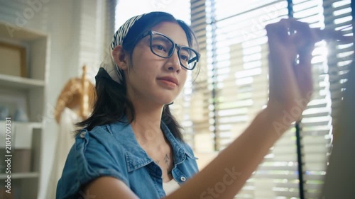 Young Asian woman artist painting on canvas easel with paintbrush in art studio, representing creative professional women designer, fine arts student lifestyle and freelance illustrator
