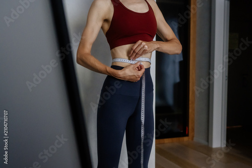 Woman wearing a sports bra and leggings measuring her waist about 60 centimeters, monitoring body progress for fitness, diet, and health goals at home