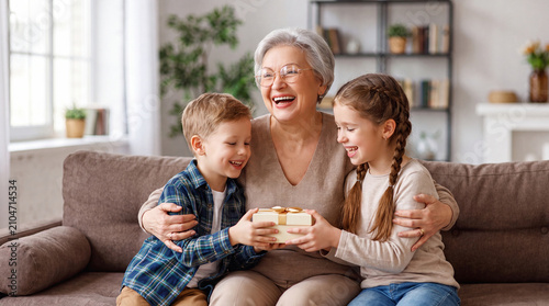 A joyful senior woman laughing as she receives a gift from her young grandson and granddaughter while sitting together on a comfortable sofa at home.