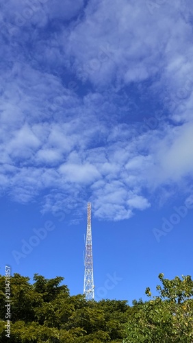 Tall communication tower above green trees under cloudy blue sky
