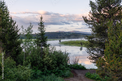 Oxbow Bend Overlook -Grand Teton National Park Wyoming.  Pines trees frame scenic view, peaceful mist shrouds a quiet Snake River at dawn, sunrise morning light, clouds reflecting in the still water. 