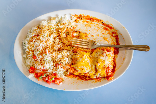 Enchilada Mexican food plate. Mexican cuisine
Partially eaten meal on white plate with fork. Rice and cheese mixed with red sauce salsa garnish. Simple, clean composition blue background