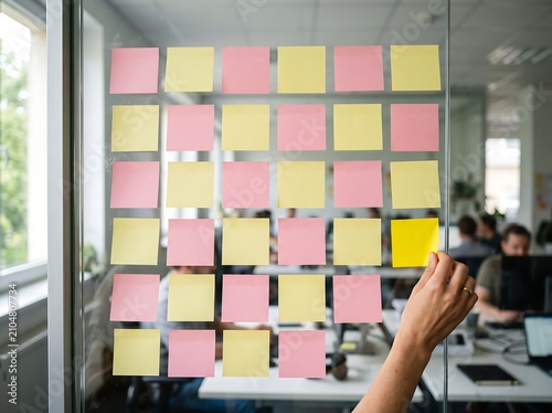 A hand places a yellow sticky note on a glass wall covered with many pink and yellow notes in an office setting.