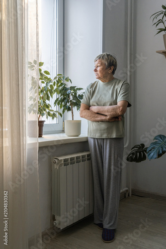 Senior woman standing by window with arms crossed, thoughtful and pensive in quiet home light, reflecting on solitude and memory