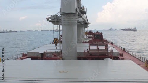 wide high angle view from a bulk carrier ship anchored at kelong anchorage in malaysia, showcasing the main deck, hatch covers, cranes and distant other anchored vessels under a bright cloudy sky