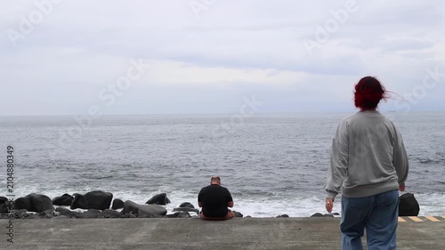 Woman in a gray sweater walking along a concrete walkway on a beach and sitting next to a man, surrounded by rocks and the ocean. Relaxed side view captures the serene and natural setting, perfect.