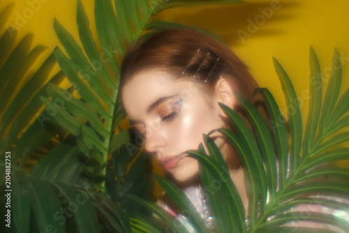 Soft portrait of a young woman surrounded by tropical leaves on a vibrant yellow background. Beauty, nature and skincare concept.