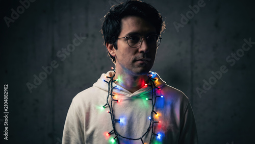 Pensive young man with glasses and a hoodie, illuminated by colorful Christmas lights.