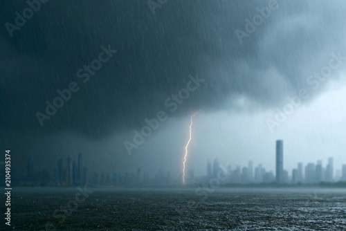 Dark clouds rage above a stormy city skyline. Single lightning bolt strikes the distant water. Rain falls heavily, obscuring the skyline. The scene evokes tension and impending danger