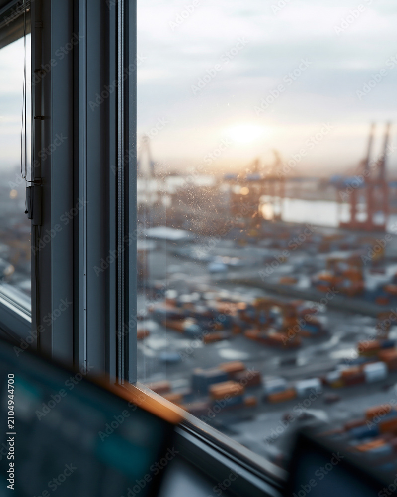 Fototapeta premium Window frames reveal a bustling port at dawn. Sunlight glows behind distant cranes and cargo containers. Raindrops streak the glass, softening the city's grit. Office screens blur in the foreground