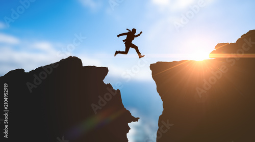 Bild auf Leinwand Businessman silhouette jumping across mountain gap during bright sunrise represe