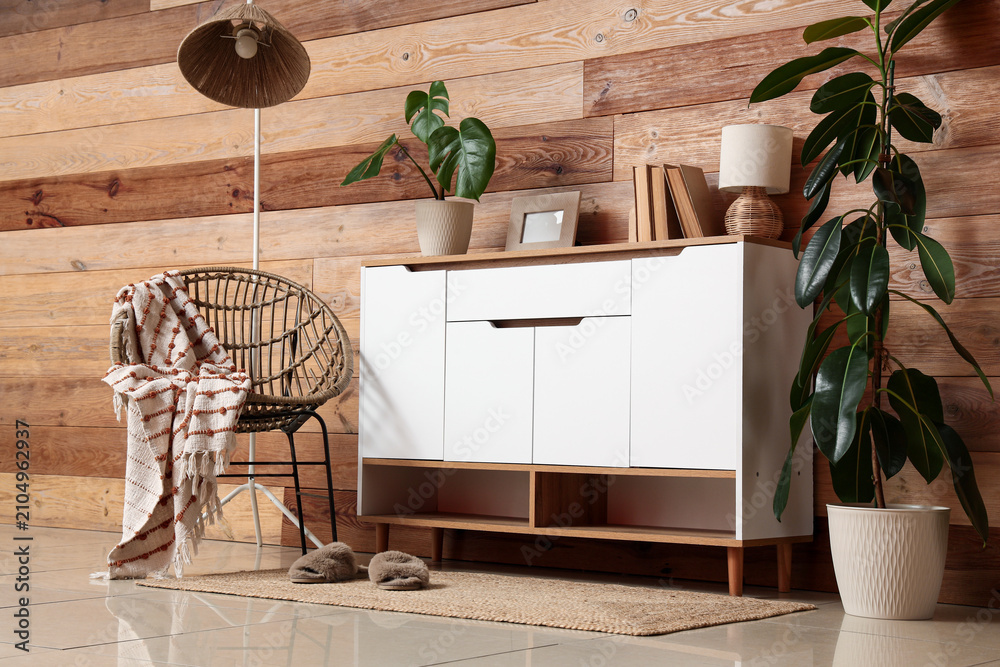 Obraz premium Cozy living room interior with wooden wall, modern white cabinet, rattan chair, and Green houseplants