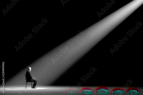 Dramatic, Minimalist Photograph of a Man Sitting Alone in a Spotlight, Evoking Solitude, Deep Thought, Performance, or the Weight of Expectation
