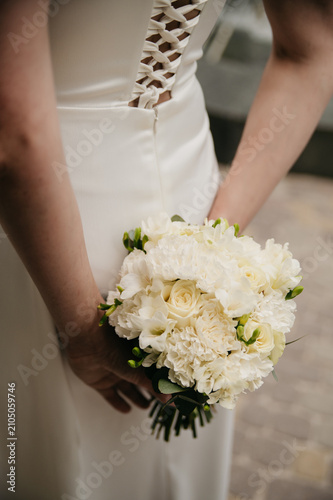 A bride in a white, tight, open dress holds a bouquet of white wedding roses behind her