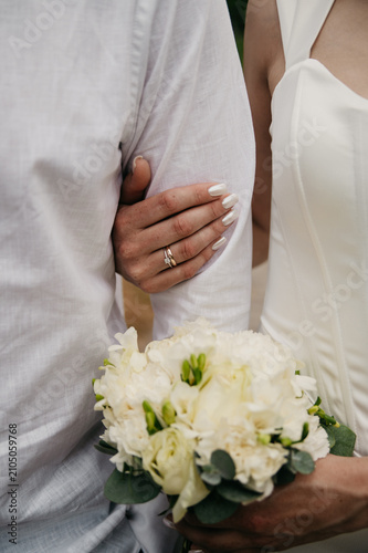 The bride in a white dress embraces her groom in a white shirt with her hand, she holds a bouquet of wedding flowers, and a gold wedding ring is worn on her fingers