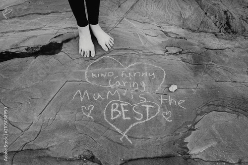 Mum is the best (kind, funny, caring). Young girl's feet standing next to a message written to her mum on rocks at the beach; Australia