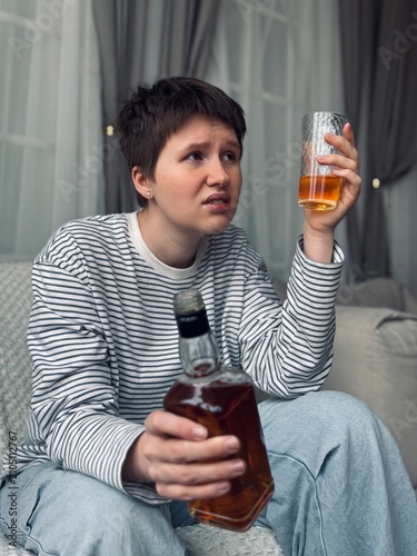 woman holding drink and bottle in indoor living room, thoughtful expression, casual attire, modern lifestyle scene, relaxing atmosphere, gray and white background, clean and natural setting.