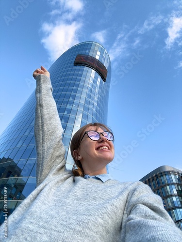Young woman raising arm for selfie beside curved glass skyscraper under bright blue sky in modern city, with woman, selfie, portrait, smiling detail, showing sunglass, urban, city, architecture
