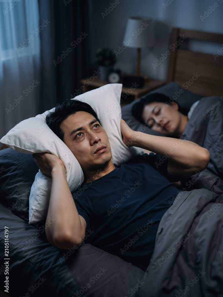 Fototapeta premium Asian man husband covering ears with pillow while wife sleeps in dark bedroom. Sleep deprivation and relationship challenges vertical banner
