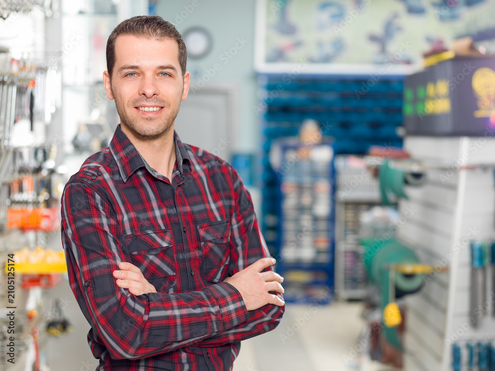 Obraz premium Portrait of a smiling male worker or store owner in a plaid shirt with crossed arms standing in a hardware store aisle next to a wall display rack full of wrenches.