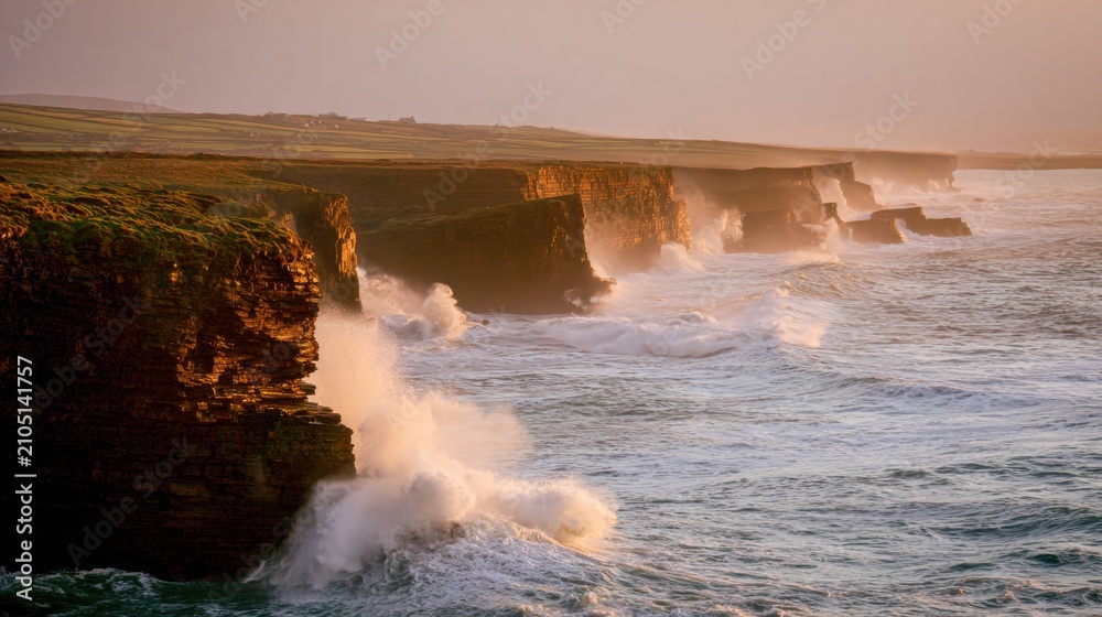 Fototapeta premium Waves crash against cliffs at sunset with a colorful sky over a coastline in Ireland