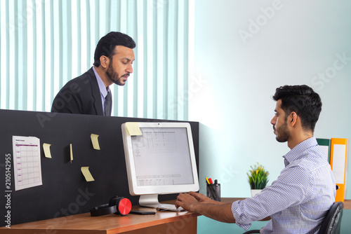 A boss in formal business suit surveying the work of subordinate employee at workdesk.