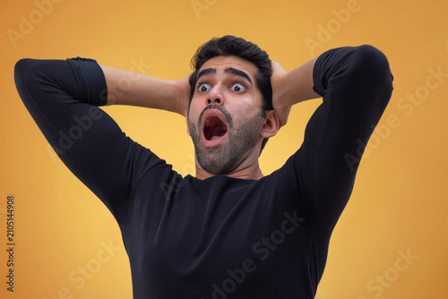 Portrait of a stunned young man with wide eyes,hands on his head and looking elsewhere with open mouth