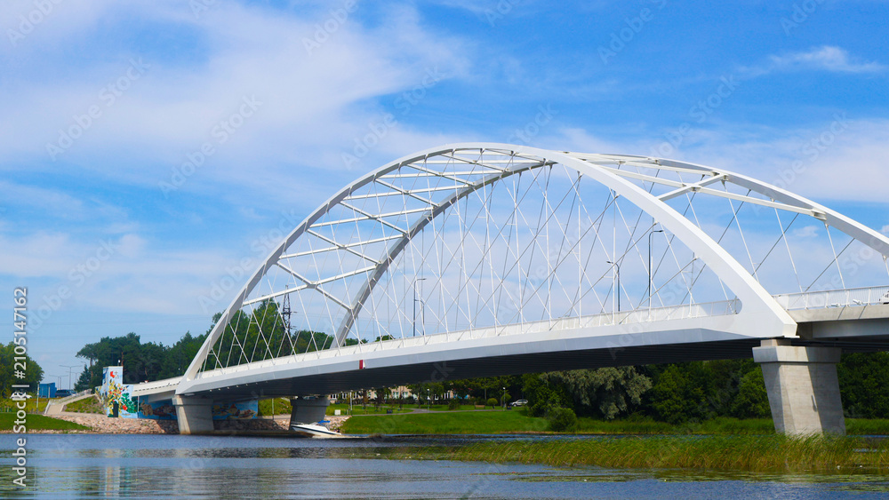 Fototapeta premium Modern white arch bridge connecting land over a reflective river with green banks, dense foliage, and a bright blue sky with wispy clouds during summer