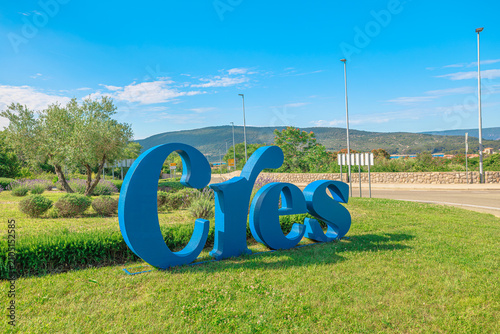 Large blue Cres sign stands on a grassy area, a landmark welcoming visitors to the Croatian island with surrounding green hills and a bright summer sky of Cres Island in Croatia