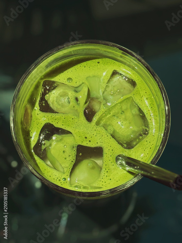 Close-up top view of vivid green iced matcha tea in clear glass with large melting ice cubes and drinking straw. Refreshing cold beverage on dark cafe table