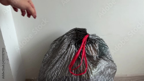 Woman's hand tightly closing a gray plastic garbage bag with red drawstring handles for proper waste disposal, getting ready for collection and recycling at home