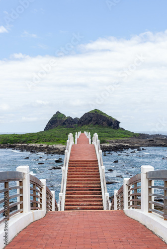 Sanxiantai bridge leading to island with green hills. Red brick pathway with white railings extending into ocean. Clear sky over coastal landscape with rocky shore.